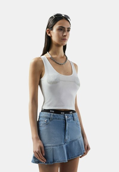Vrouw draagt een wit cropped tanktop met parelversieringen, een blauwe denim minirok met ruches, een zilveren ketting en een zonnebril op haar hoofd.