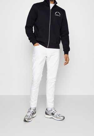 Sudadera navy con cremallera y logo blanco, pantalones blancos y zapatillas deportivas plateadas. La tela parece suave, con un cuello que se levanta y bolsillos.