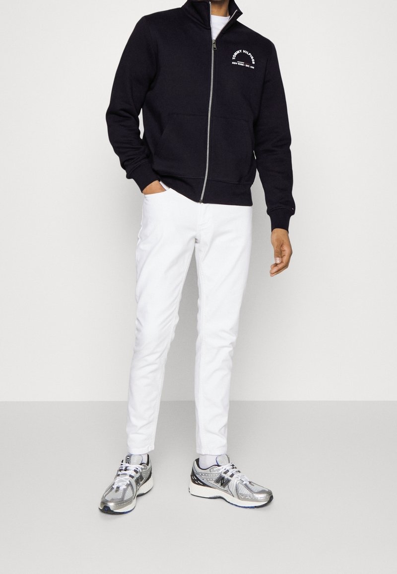 Felpa navy con zip e logo bianco, pantaloni bianchi e scarpe sportive argentate. Il tessuto appare liscio, con colletto alto e tasche.