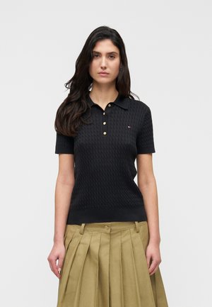 Femme portant un polo noir texturé à manches courtes avec des boutons dorés et une jupe plissée beige, debout devant un fond uni.
