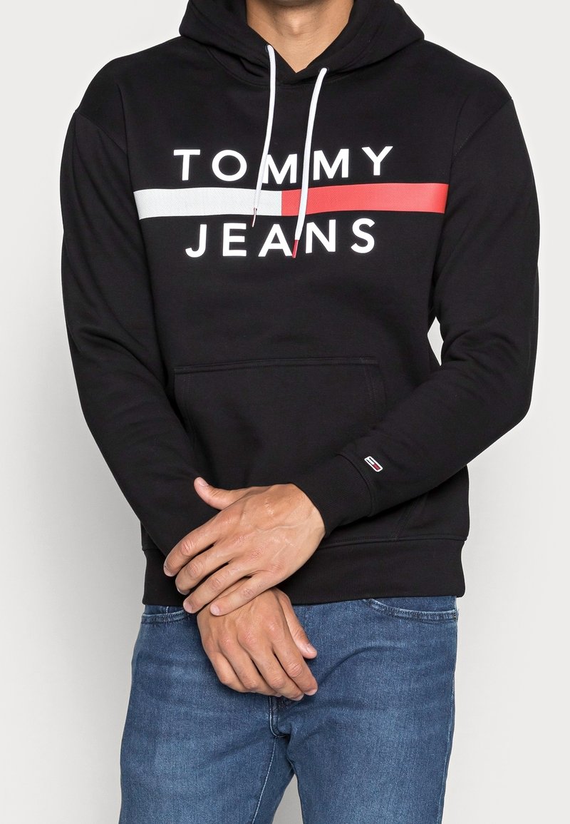 Homme portant un sweat à capuche noir avec le logo "Tommy Jeans" blanc et rouge, cordons blancs, et un jean bleu, les mains jointes devant.