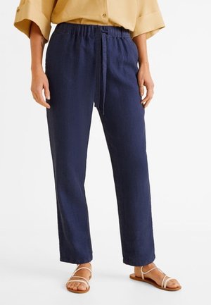 Kobieta w granatowych lnianych spodniach z troczkami, beżowej koszuli na guziki i białych płaskich sandałach na paski stoi na tle białego tła.