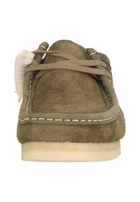Olivgrön mockasinliknande sko i mocka med vit sömnad, rund tå och beige snörning. Har en platt gummisula och en mjuk textur.