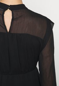 Blouse noire en tissu transparent plissé, avec une ouverture en forme de trou de serrure dans le dos et une fermeture par bouton au niveau du col.
