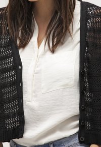 Cárdigan negro de punto abierto sobre una camisa blanca con botón y un bolsillo en el pecho. Diseño con cuello, mangas cortas y detalles en la tela texturizada.