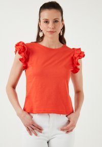 Raudona trumpomis rankovėmis palaidinė su banguotu akcentu ant pečių; pagaminta iš minkštos medžiagos; derinama su baltais kelnėmis. Modelis pozuoja su rankomis ant klubų.