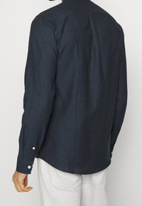 Camicia a maniche lunghe blu scuro in tessuto testurizzato, con polsini abbottonati e vestibilità rilassata. Presenta una piega sul retro per facilitare i movimenti.