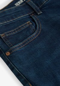 Donkerblauwe spijkerbroek met een close-up van de voorzak, gekenmerkt door koperen stiksels en een metalen knoop. Getextureerde stof met een lichte vervaging.