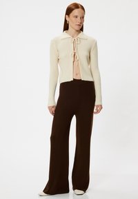 Crème gebreide cardigan met ribbelstructuur, een kraag en een sluiting met voorste strik, gecombineerd met losse bruine flared broeken en witte schoenen met puntige neuzen.
