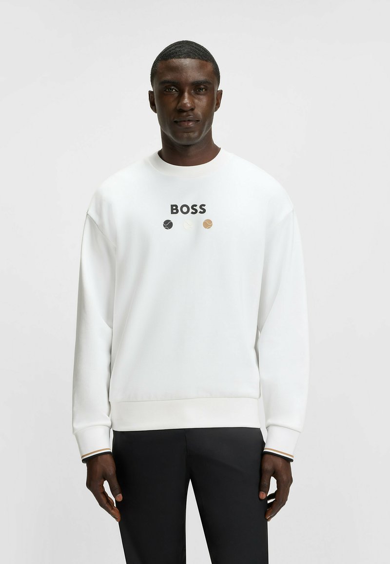 Felpa bianca con logo "BOSS" in nero sopra tre cerchi colorati. Presenta un collo a giro e polsini a costine. Abbinata a pantaloni neri.