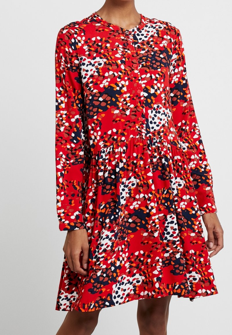 Femme portant une robe rouge à manches longues avec un motif abstrait multicolore à pois et un empiècement avant boutonné.