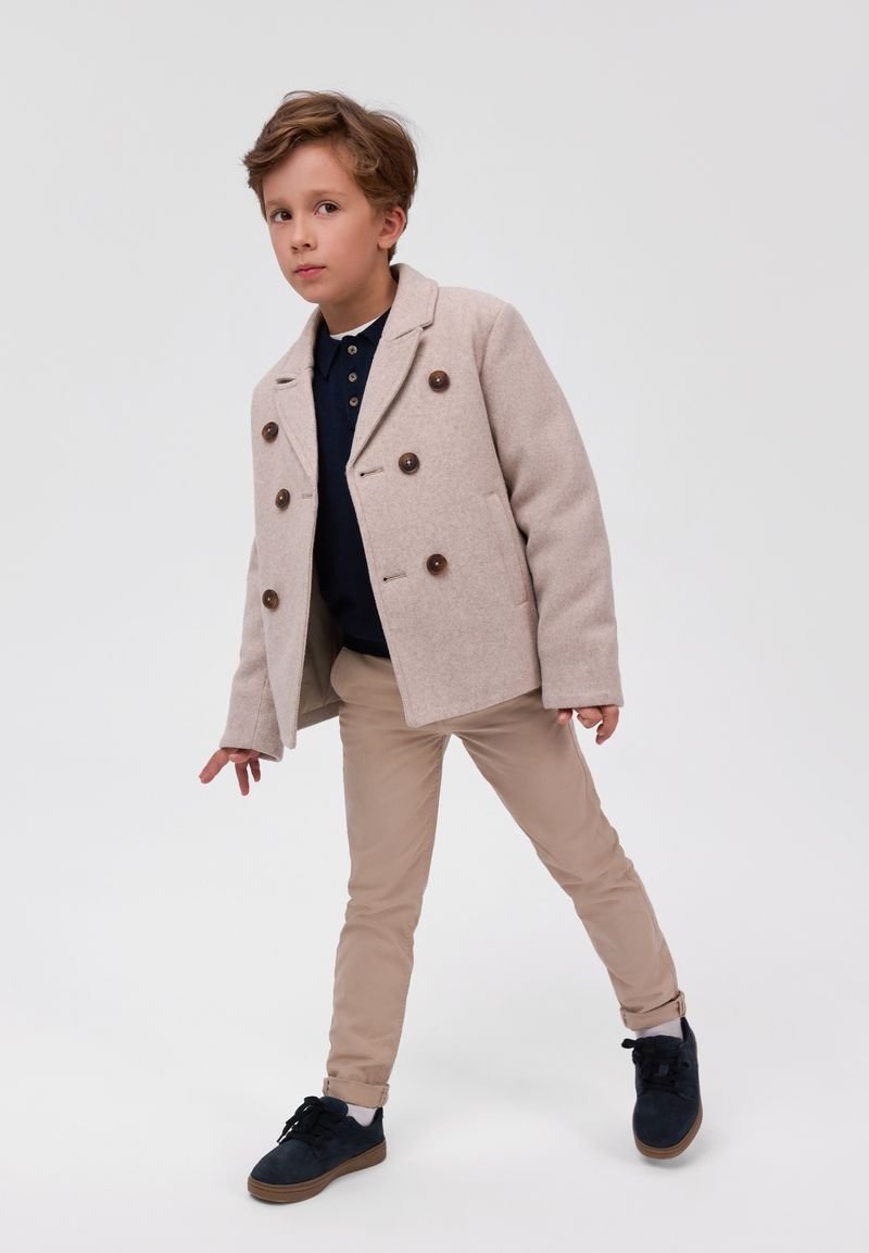 Jeune garçon portant un manteau beige croisé, une chemise bleu marine, un pantalon beige et des chaussures noires, debout devant un fond clair uni.