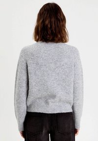 Personne avec des cheveux bruns mi-longs portant un pull en maille gris clair et un jean noir, se tenant de dos contre un fond uni.