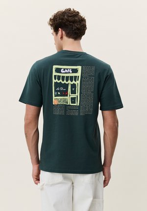 Mann trägt dunkelgrünes T-Shirt mit Café-Schaufenster-Grafik und Text auf dem Rücken, kombiniert mit weißer Hose, vom Kamera weggewandt.