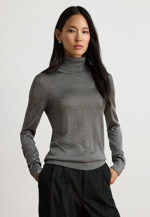 Frau mit langen dunklen Haaren, die einen taillierten grauen Rollkragenpullover und dunkle Nadelstreifenhosen trägt, steht mit den Händen in den Taschen.