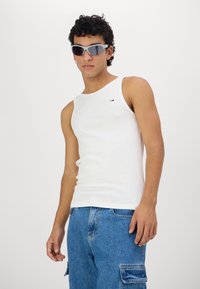 Fehér pamut tank top kerek nyakkivágással és kis logóval. Kék farmer cargo rövidnadrággal párosítva, ami oldalzsebekkel rendelkezik. A modellen napszemüveg van.