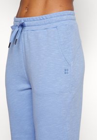 Ljusblå atletiska joggers i mjukt tyg, med elastisk midja och snörning samt sidofickor. Liten logotyp synlig.