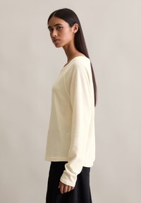 Long-sleeve, light yellow top made of textured fabric, featuring a boat neckline and extended cuffs, paired with dark trousers.
