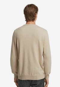 Man med brunt hår klädd i en beige tröja och svarta jeans, stående vänd bort mot en plain vit bakgrund.