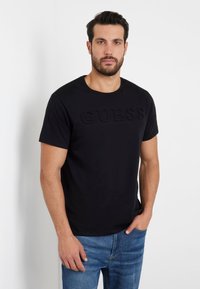 T-shirt in cotone nero con logo "GUESS" in rilievo sul davanti. Collo a giro, maniche corte, abbinato a jeans in denim blu per creare un contrasto.