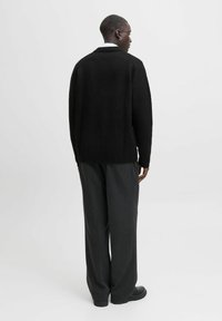 Melna adīta džemperis ar brīvu piegriezumu, ar ribotām apakšmalu un piedurknēm, apvienots ar tumšajiem pelēkajiem biksēm, skatīts no aizmugures.