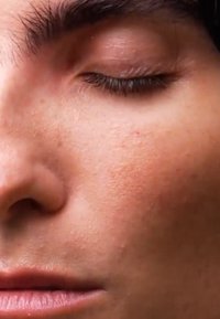 Nærbilde av et ansikt med glatt, solbrun hud, lukkede øyelokk, subtile fregner på kinnet og lys ansiktshår nær leppene.