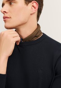 Maglione navy a maglia con collo rotondo, caratterizzato da un piccolo logo in rilievo. Sciarpa marrone indossata sotto, con texture liscia e colore scuro.