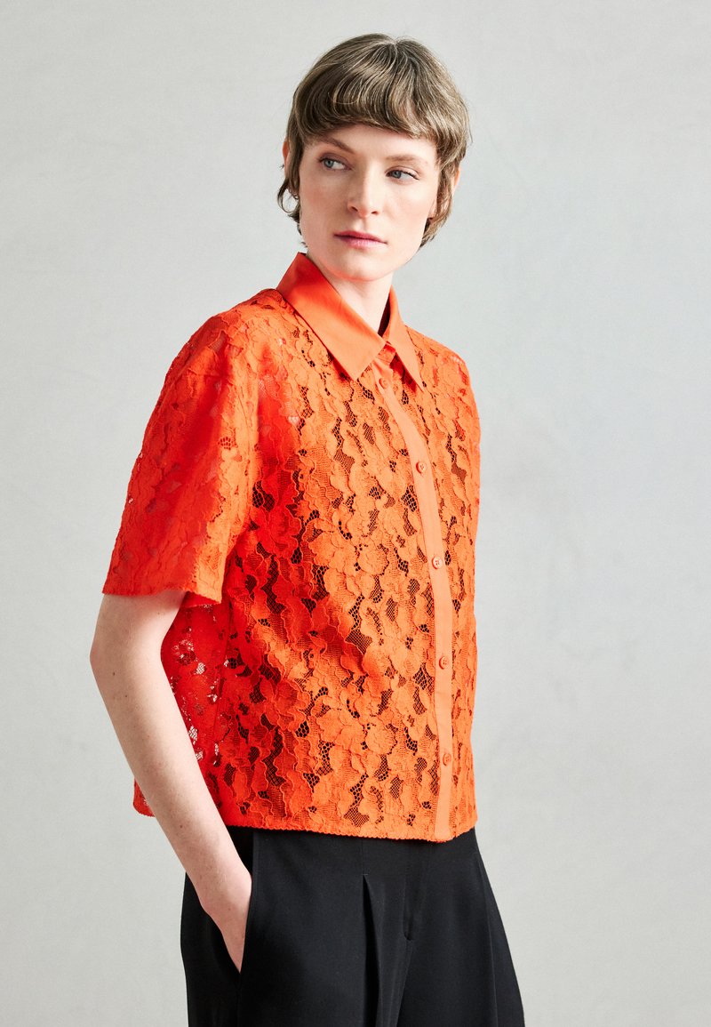Oranje korte mouw kanten shirt met een knoopsluiting aan de voorkant, bloemmotief en klassieke kraag, gecombineerd met een zwarte broek.