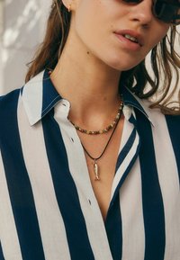 Chemise rayée bleu marine et blanche avec détails de boutons. Porte un collier de perles multicolores et un pendentif en forme de poisson sur un cordon noir.