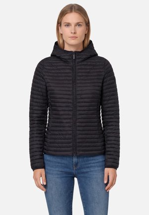 Vrouw die een zwart doorgestikt gewatteerd jack met capuchon en ritssluiting aan de voorkant draagt, gecombineerd met een blauwe spijkerbroek, staand tegen een effen lichte achtergrond.