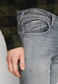 Jeans de mezclilla gris con un corte ajustado, que presentan bolsillos frontales y un botón de metal. Mano descansando sobre el muslo. Camisa a cuadros verde oscuro visible.
