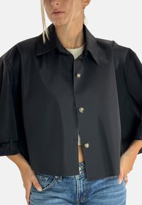 Schwarze, kurz geschnittene Jacke mit Kragen, großen Ärmeln und glänzenden Knopfverschlüssen, kombiniert mit einem hellen Oberteil und Jeans.