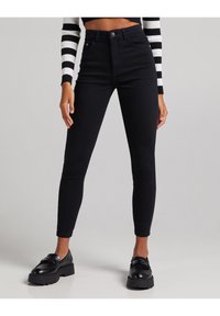 Černé džíny s vyšším pasem a úzkým střihem, se zapínáním na knoflík, pěti kapsami a jemným prošíváním. Hladká textura, bez vzorů.