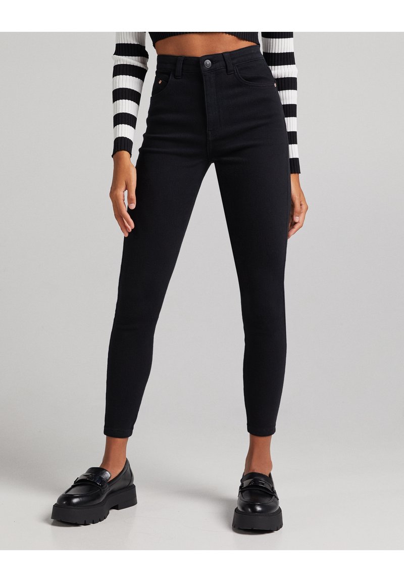 Černé džíny s vyšším pasem a úzkým střihem, se zapínáním na knoflík, pěti kapsami a jemným prošíváním. Hladká textura, bez vzorů.