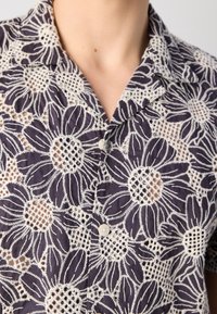 Gros plan d'une chemise bleu marine avec une grande broderie en dentelle florale blanche et une fermeture à boutons, montrant l'encolure et le haut de la poitrine.