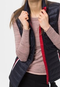 Mörkblå quiltad väst med röd foder och vita detaljer, med dragkedjestängning och sidofickor, ihop med en ljusrosa lång ärmad topp.