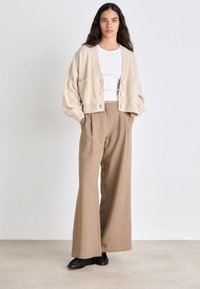 Beige gebreide cardigan met een cropped ontwerp, voorzien van grote knopen en zijzakken. Gecombineerd met hooggeplaatste, wijde bruine broeken.