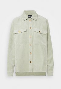 Ljust grön kordflorskjorta med krage, två bröstfickor och bruna knappar; strukturerad tyg med subtila vertikala ränder.