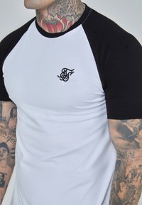 Camiseta blanca con mangas raglán negras, que presenta un pequeño logotipo negro bordado en el pecho. La tela tiene una textura suave.