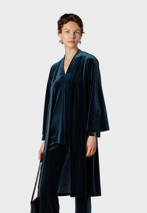 Femme aux cheveux courts et bouclés, portant un manteau long en velours bleu foncé et un pantalon assorti, tenant un sac noir sur un fond blanc.
