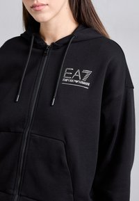 Crna zip-up dukserica napravljena od mekanog materijala, s kenguru džepom, kapuljačom s vezicom i EA7 Emporio Armani logom u srebrnoj boji.