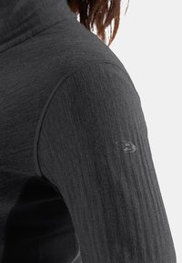 Top de manga larga negro con cuello alto, que presenta un diseño de punto texturizado y un logotipo bordado en el hombro. Acabado de tela suave.