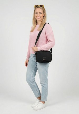 Lächelnde Frau mit einem rosa Pullover, hellblauen Jeans, weißen Sneakers, gelber Sonnenbrille auf dem Kopf und einer schwarzen Umhängetasche.