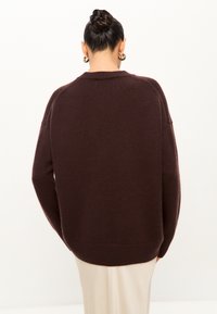 Pulover tricotat gros, de culoare maro, cu o croială relaxată, umeri lăsați și margine cu nervuri. Textură moale și design simplu, potrivit pentru stratificare.