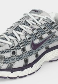 Λευκό και γκρι παπούτσι τρεξίματος Nike με μοβ σήμα, διχτυωτά πάνελ, λευκά κορδόνια και παχιά υφασμένη σόλα σε κοντινό πλάνο.