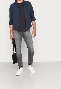 Laivaston vetoketjuhuppari mustan t-paidan päällä, yhdistettynä harmaisiin farkkuihin ja valkoisiin lenkkareihin. Henkilö pitää mustaa olkalaukkua.