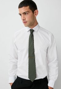 White long-sleeve shirt paired with a dark green patterned tie, featuring a sleek gold tie clip; worn with black trousers.