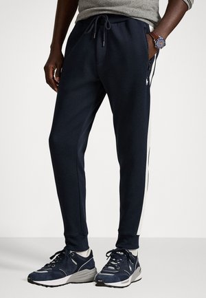 Granatowe dresy z elastycznym paskiem, boczną kieszenią na zamek, białymi paskami po bokach i ściągaczami na kostkach, w połączeniu z szarymi trampkami.