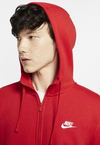 Rode Nike zip-up hoodie met een trekkoordcapuchon, wit logo op de borst, gladde stoftextuur en een aansluitende pasvorm.