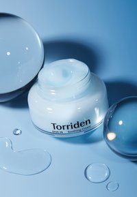 Torriden DIVE IN Rauhoittava Kerma kirkkaassa purkissa, vaaleansininen kerma sisällä, ympärillä kiiltäviä sinisiä palloja ja pisara sinisellä pinnalla.